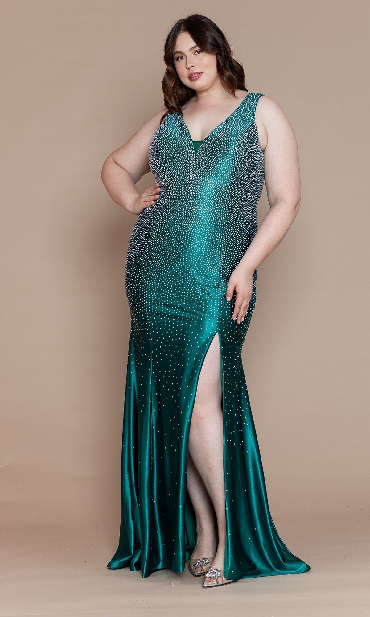 A woman stands against a beige background wearing the Spandex Long Beaded Plus-Size Prom Dress W1130, a sleeveless, emerald green, floor-length gown featuring rhinestone accents and a thigh-high slit.